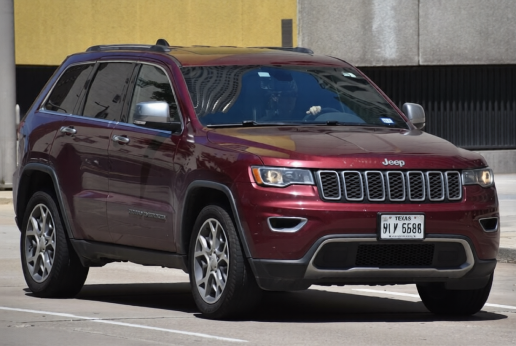 A portrait of a maroon-colored Jeep Grand Cherokee traveling down a highway in moderate traffic