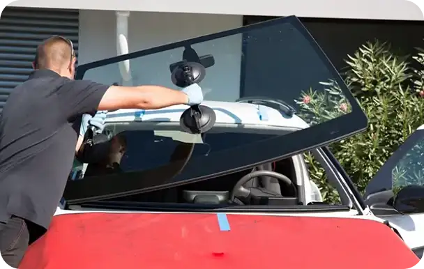 Auto glass technician installing a new windshield on a red car using suction cup tools, performing the job outdoors near a repair facility.