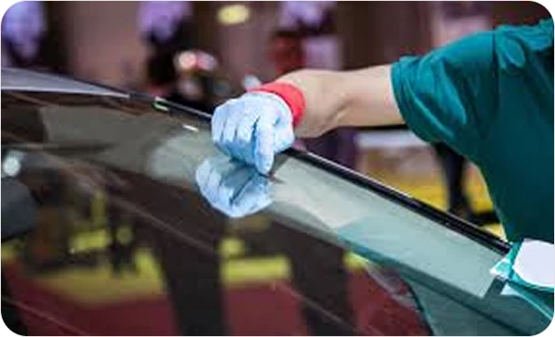 A gloved auto glass technician carefully positions or installs a car windshield, holding the top edge of the glass during the replacement process.