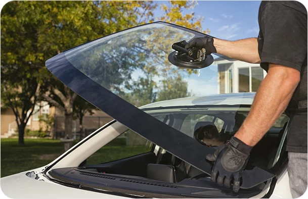 Technician wearing gloves uses a suction cup to install a new windshield on a white car outdoors.
