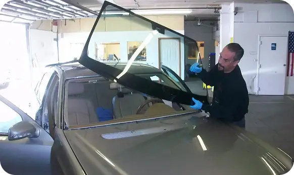 Technician carefully installing a new windshield on a car inside an auto glass repair shop, wearing safety gloves and using professional tools.