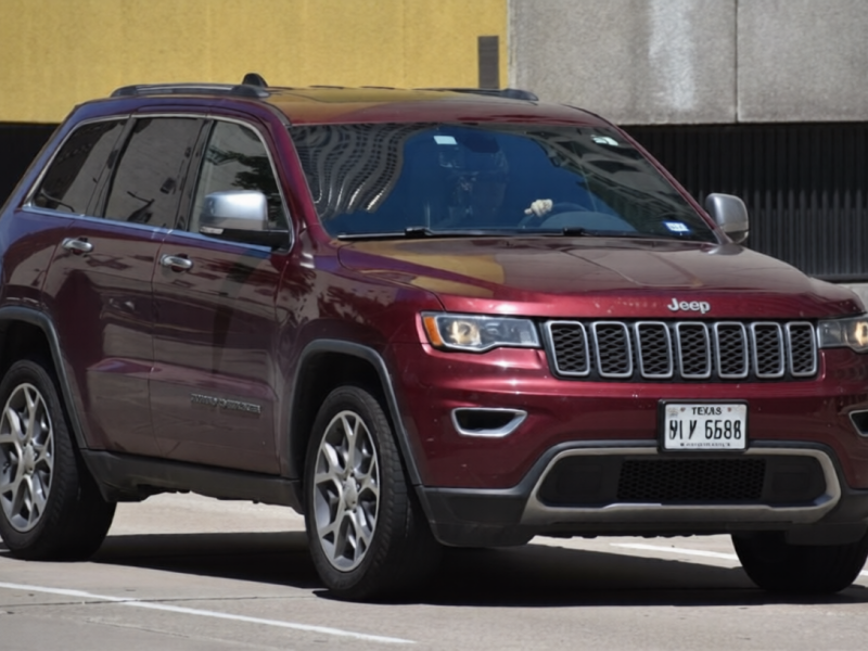 A portrait of a maroon-colored Jeep Grand Cherokee traveling down a highway in moderate traffic