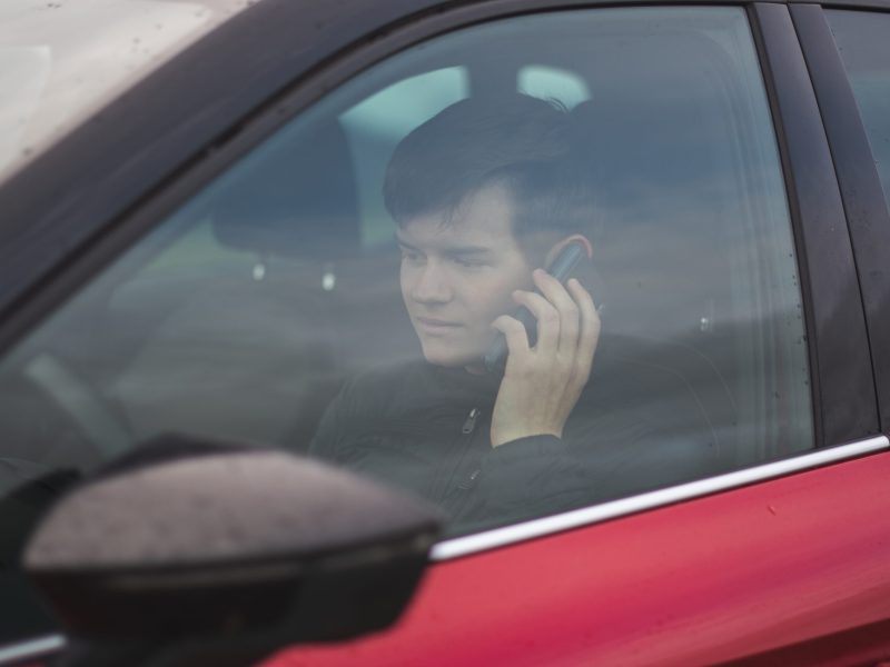 a view of a young male wearing a black jacket sitting in a red car while talking on the phone