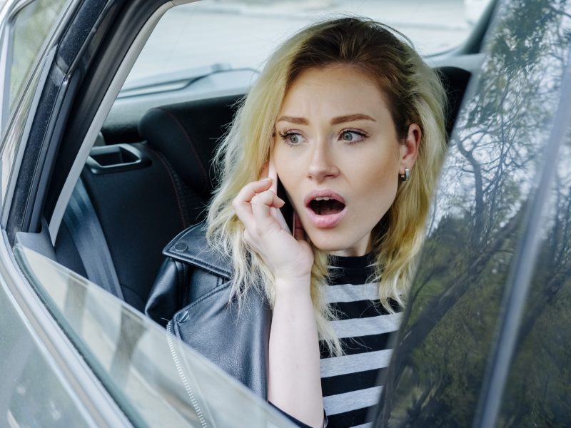 young surprised woman talking on smartphone while travelling in the back seat of car wearing striped top and black leather jacket